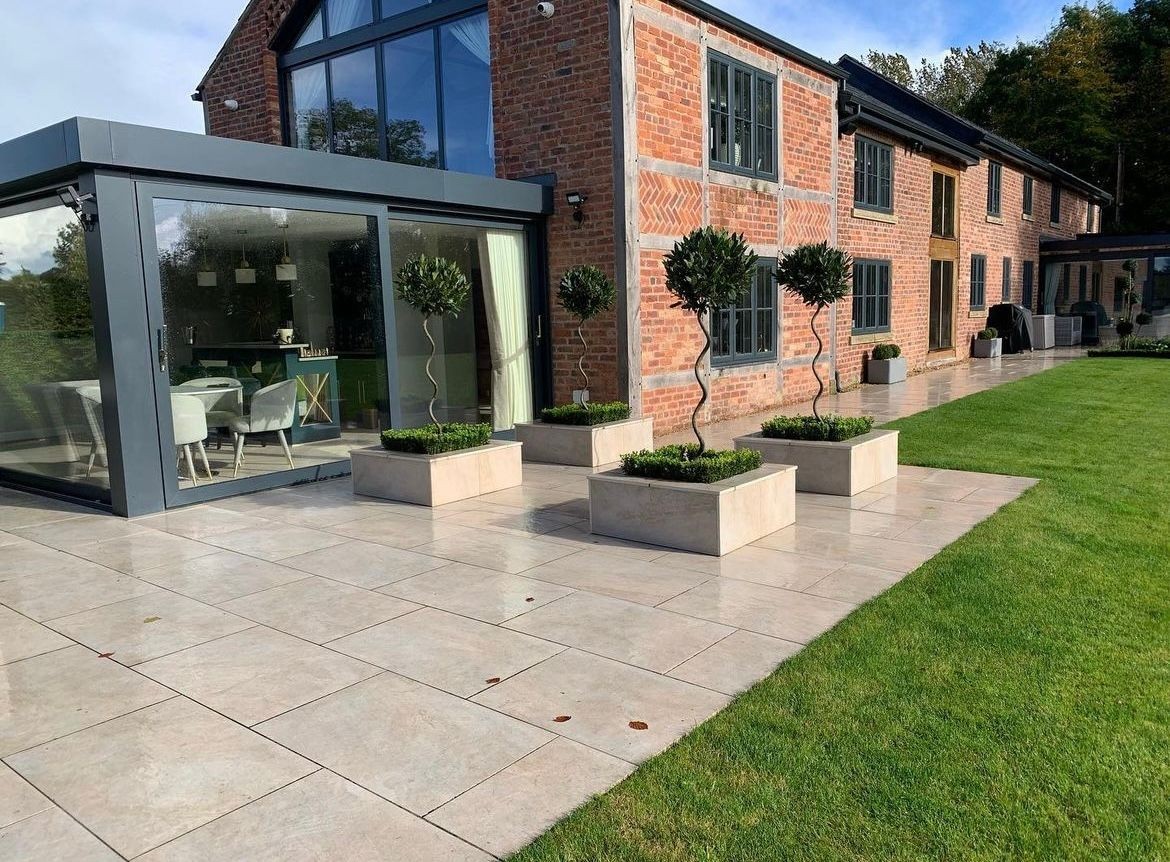 Modern patio with potted trees, tiled flooring, and glass extension on a brick house.