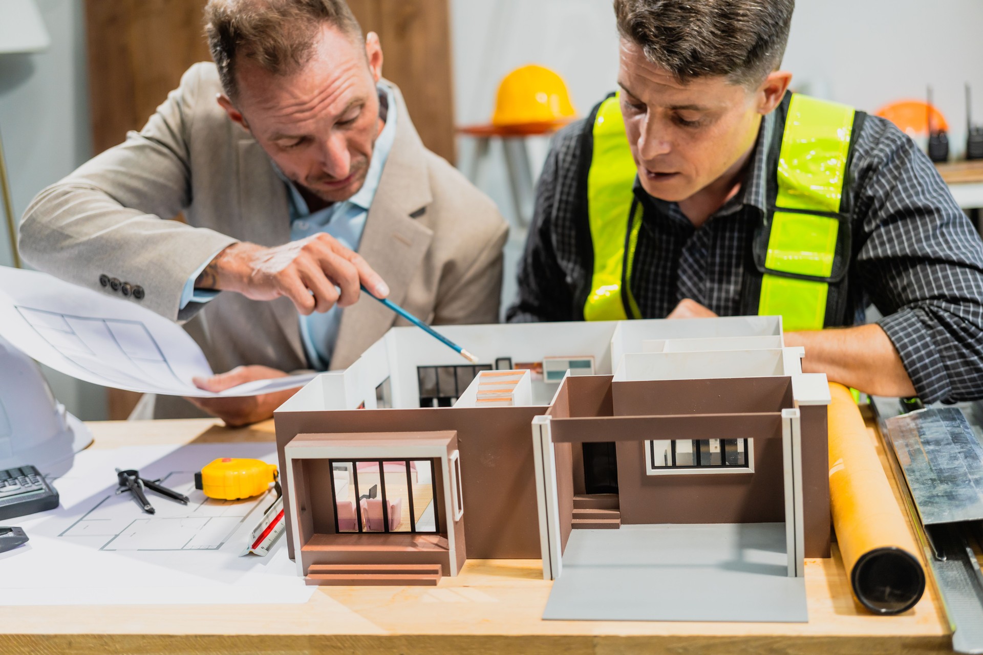 engineer contractor and an Italian architect engineer are seated at a desk, discussing renovation plans with blueprints and a house model in front of them.